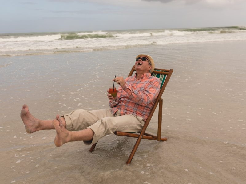 Side view of active senior man having cocktail drink while relaxing in a sun lounger at the beach with ocean in the background. He wears a hat